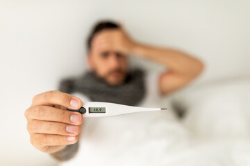 Fever. Ill man showing thermometer with high temperature result to camera, lying on bed. Selective focus