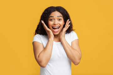 Wow offer. Overjoyed african american lady looking at camera in excitement and touching face, yellow studio background