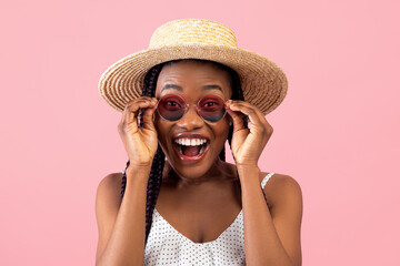 Excited millennial African American woman in summer wear touching her sunglasses, shouting WOW over pink background