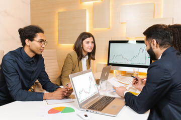 Millennial colleagues sitting at office desk, looking at computer and talking about business issues. Diverse employees communicate near each other and negotiating on projects together