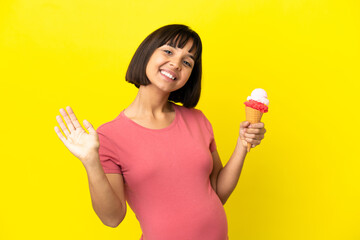 Pregnant woman holding a cornet ice cream isolated on yellow background saluting with hand with happy expression