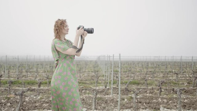Young pretty woman takes pictures in harvest field. Action. Side view of female photographer in a field on grey sky background.