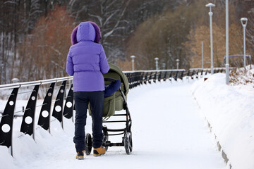 Girl walking with a baby stroller in winter park. Snow weather, concept of motherhood, single mom...