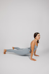 Fototapeta premium Brunette poses on isolated background. Young woman in gray tracksuit performs exercise from yoga or stretching, she stands in cobra pose and smiles, side view. 