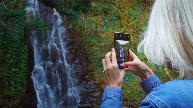 A young woman holds a smartphone and takes photos standing near a high waterfall with foaming streams of water, rear view. She is wearing a denim jacket, Makhuntseti Georgia.