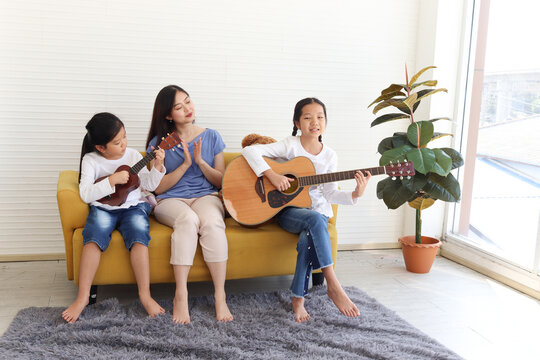 Two happy daughter girls and mother singing a song and playing guitar ukulele while sitting on sofa in living room, mom and children spending special moment together.