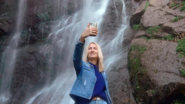 A young woman holds a smartphone and takes pictures of herself standing near a high waterfall with foaming streams of water. She's wearing a denim jacket. Selfie. George's Makhuntseti.