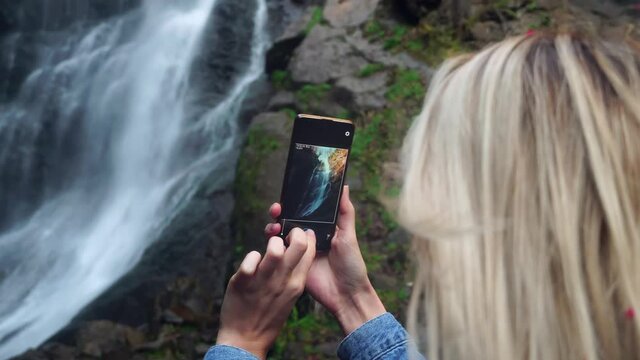 A young woman holds a smartphone and takes photos standing near a high waterfall with foaming streams of water, rear view. She is wearing a denim jacket, Makhuntseti Georgia.