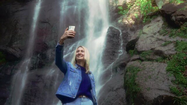 A young woman holds a smartphone and takes pictures of herself standing near a high waterfall with foaming streams of water. She's wearing a denim jacket. Selfie. George's Makhuntseti.