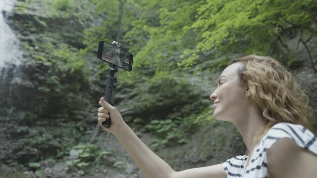 Woman taking pictures or shooting video of mountains and waterfall on her smartphone using a tripod. Action. Female traveler with a selfie stick and a phone standing in a gorge surrounded by green