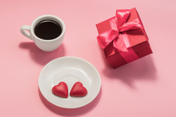 Obraz na płótnie Canvas A cup of coffee, two chocolate hearts on a saucer and a gift box on a pink background. Love, birthday, engagement and Valentine's Day concept. Festive background.