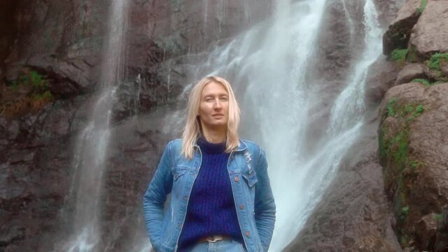A young woman pulls a smartphone out of her pants pocket near a high waterfall with foaming streams of water. She's wearing a denim jacket. Selfie. George's Mahuntseti. slow motion,