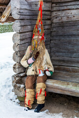 Doll in Meteņi mumming mask and costume sitting in a bench near an old wooden house. Meteni is ancient Latvian spring waiting holiday, when mummers scare the winter.