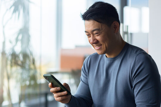 Close-up photo of male businessman using phone in modern office, asian looking at phone display and smiling, online call