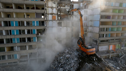 Excavator for dismantling buildings, using a special claw, dismantles a multi-storey building. The sun's rays break through a thick cloud of dust in front of the ruined building