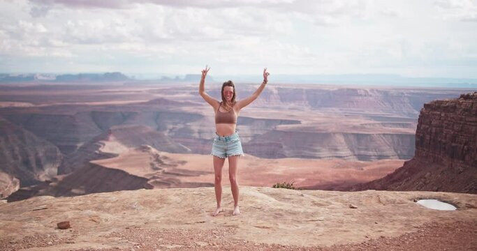 Beautiful young female model dancing and waving peace signs on the edge of a cliff at Goosenecks State Park in the desert of Utah