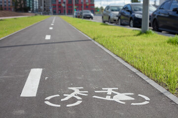 Obraz premium New empty bicycle path closeup with green grass and cars around. White bike symbol on road. Bicycle infrastructure in the city. Bikeway with no people. Separated road for cycling.