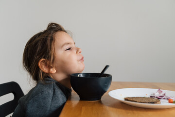 Unhappy child girl eats soup from black bowl with bread and onion. Lifestyle photo of kid in...