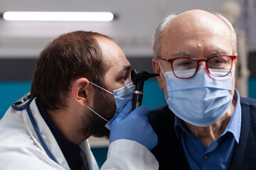 Medic using otoscope to do ear checkup on senior man, doing otolaryngology consultation with patient. Doctor with face mask examining infection to give prescription treatment. Close up