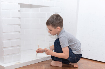 Naklejka premium Child boy sits on the floor and paints a decorative fireplace with white paint gouache. Home improvement, DIY concept. Selective focus