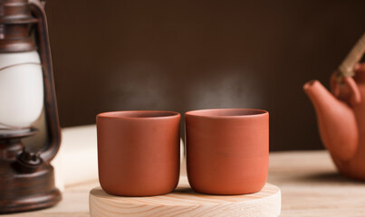 Two cups with hot steamed brown clay tea or coffee, on a wooden table with partial view of the lamp and the kettle or coffee pot. brown background. Space for text.