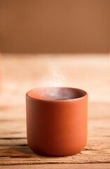 Cup of tea or coffee with hot steam from brown clay, on rustic wooden table, brown background