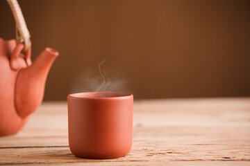 Brown ceramic mug with tea or coffee and hot steam, on rustic wooden table, partial view of teapot or coffee pot, brown background, space for text.