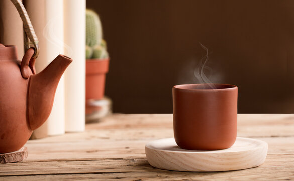Cup of tea or coffee with hot steam from brown clay, on wooden table with partial view of teapot or coffee pot. books, cactus. Brown background.