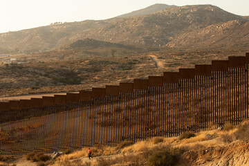 Tecate, Baja California, Mexico - September 14, 2021: Late afternoon sun shines on the USA Mexico border wall people walk in front of it. - obrazy, fototapety, plakaty