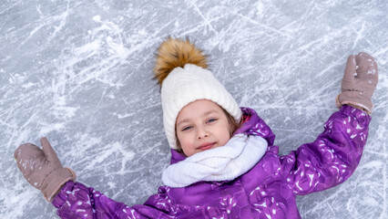 A cute little girl, in a white hat and a purple jacket, lying on the ice at the skating rink, in...