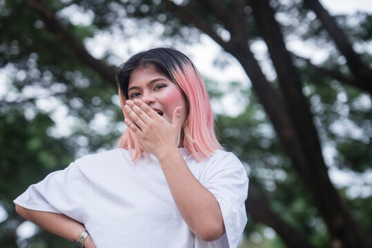 A happy young asian woman with pink highlights pretends to yawn showing boredom or an expression of being unimpressed. Outdoor scene.