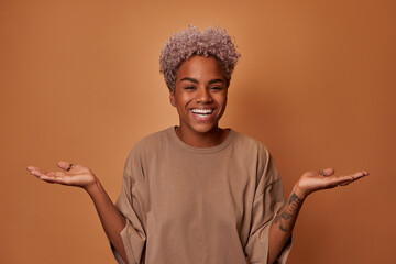Surprised joyful young African American woman looks directly at you smiling broadly spreading hands, demonstrating incomprehension and indecision standing in casual clothes on brown background - obrazy, fototapety, plakaty