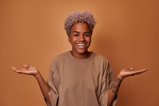 Surprised joyful young African American woman looks directly at you smiling broadly spreading hands, demonstrating incomprehension and indecision standing in casual clothes on brown background
