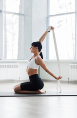 Girl in a white top and black shorts and bicycles doing yoga sitting on a black gym Mat training her hands with an elastic band on a white background. Morning exercise