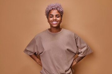Young attractive ethnic African American woman curly hairstyle smiles broadly and looks at camera holds hands on belt posing standing in beige oversized t-shirt on brown studio background