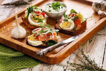 Appetizing canapes or sandwiches with smoked salmon. Toast with fish, soft cheese, eggs, onion rings and salad. Delicious breakfast or snack on a wooden tray. Food still life. Copy space