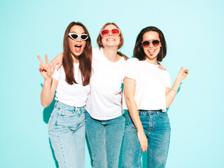 Three young beautiful smiling hipster female in trendy same summer white t-shirt and jeans clothes. Sexy carefree women posing near light blue wall in studio. Cheerful and positive models having fun