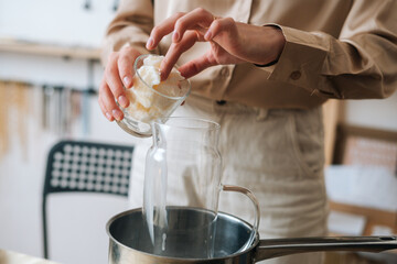 Close-up cropped shot of female artisan adding white dry soy wax in glass jar into pot of boiling...
