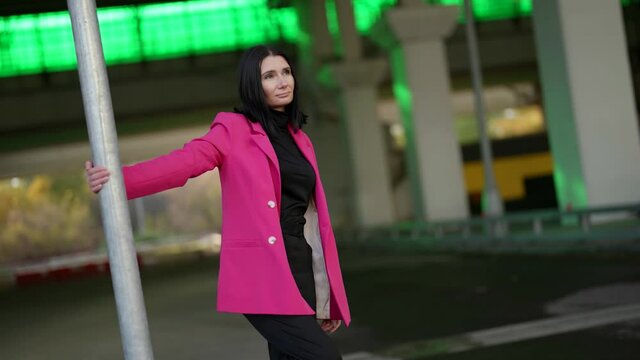 a brunette in a bright pink coat thoughtfully holds hand to a pole against the background of green glowing windows of a moder building