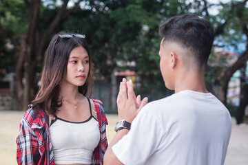 A young asian man pleads with his pokerfaced girlfriend, who is avoiding eye contact, for...