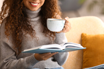 Young African-American woman with cup of tea reading book on sofa at home