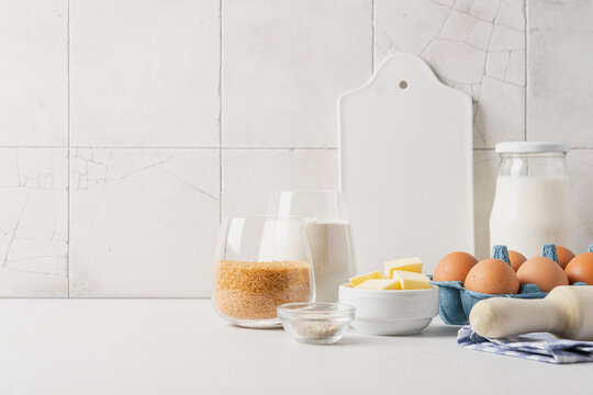 Variety of ingredients and baking utensils on white table with ceramic tile wall in the background. Kitchen background with copy space