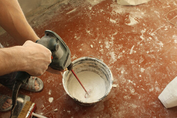 Close-up of a builder's hands holding a drill with a nozzle stirring a putty solution in a bucket....