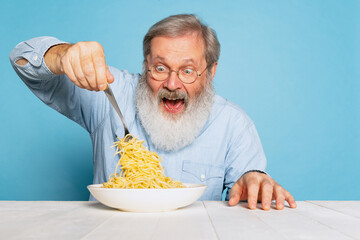 Excited senior hairy gray-bearded man eating large portion of noodles, pasta isolated on blue...