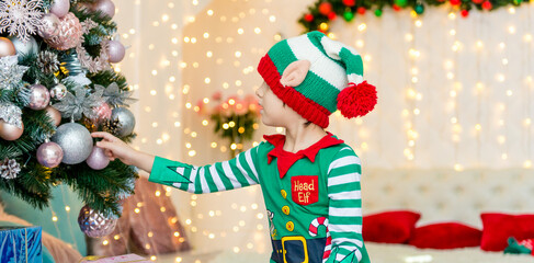 Child decorating Christmas tree at home. Little boy in elf costume and hat with Xmas ornament. Family with kids celebrate winter holidays. Kids decorate living room and fireplace for Christmas.