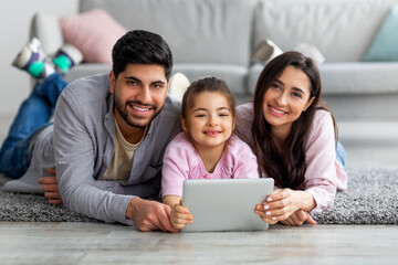 Young arab parents and their cute daughter using digital tablet while lying on floor together and smiling to camera