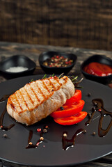 Steak with sliced tomatoes in a black plate. Meat on a dark wooden background. Food, sauce, salt, pepper and spices on an old shabby table.