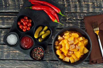 Boiled potatoes with meat in a black bowl. Seasonings and spices on a black wooden table. Ketchup, salt, pepper on an old shabby background.