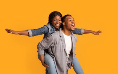 Joyful Black Couple Having Fun Together Over Yellow Background, Woman Piggybacking Boyfriend