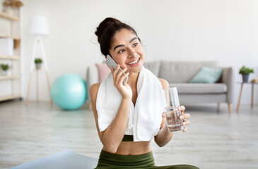 Fit young Indian woman holding glass of water, talking on smartphone, having break from domestic training